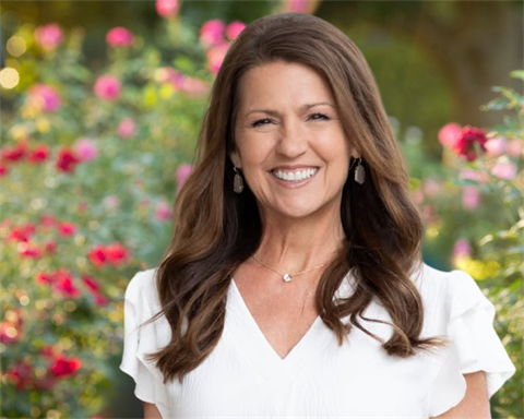 White woman with dark hair in a white shirt smiling with roses behind her.