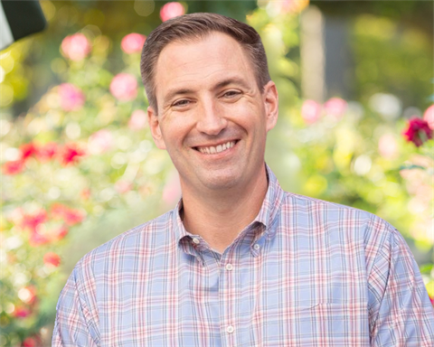 White male smiling in a blue plaid shirt with roses behind him.